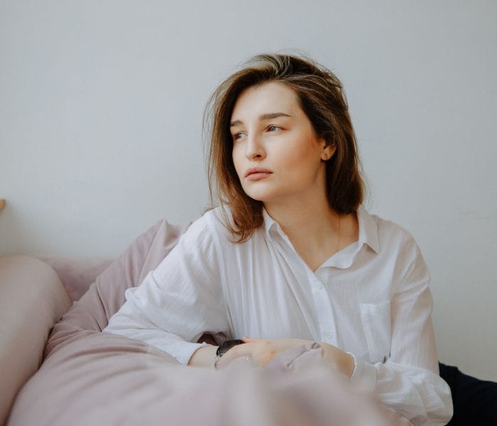 A person seated on a bed, resting their arms on a pillow while looking out of frame in a quiet indoor setting.