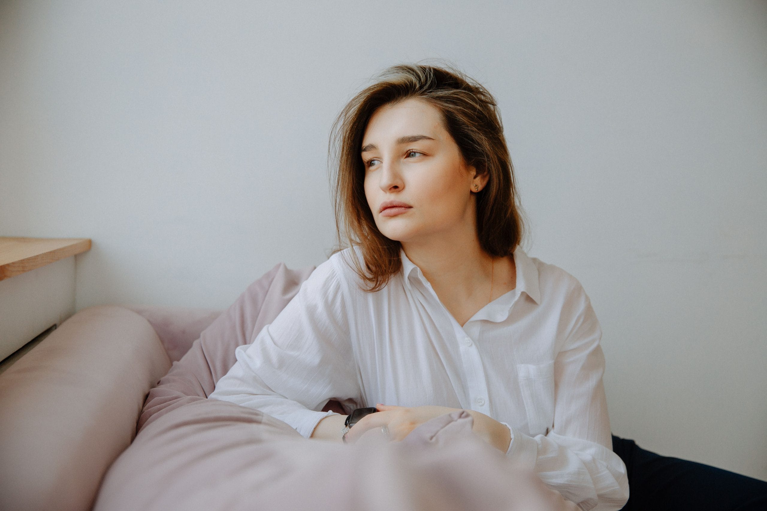 A person seated on a bed, resting their arms on a pillow while looking out of frame in a quiet indoor setting.