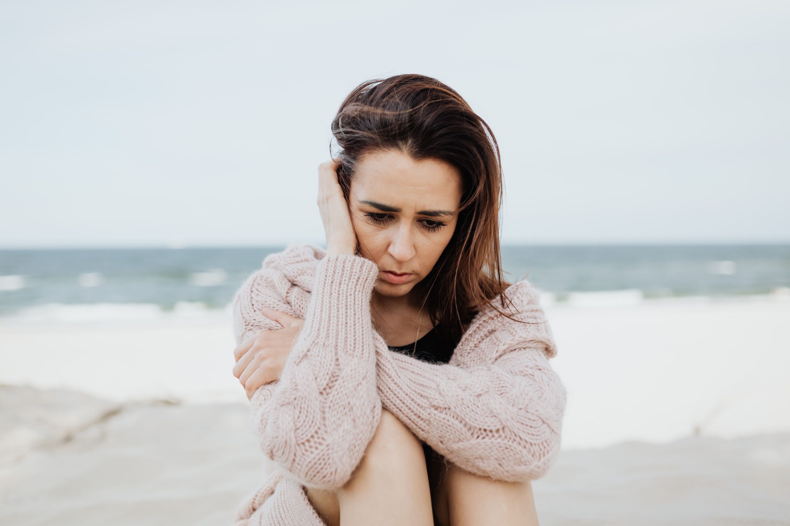 Person sitting alone on a beach, embracing themselves while appearing deep in thought and emotional distress.