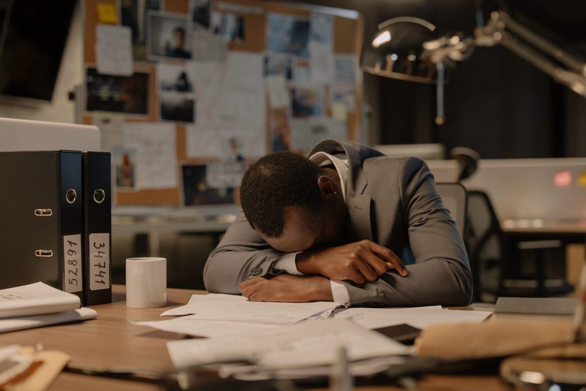 A tired professional resting his head on his arms at a desk surrounded by paperwork, showing signs of work-related stress and overwhelm.