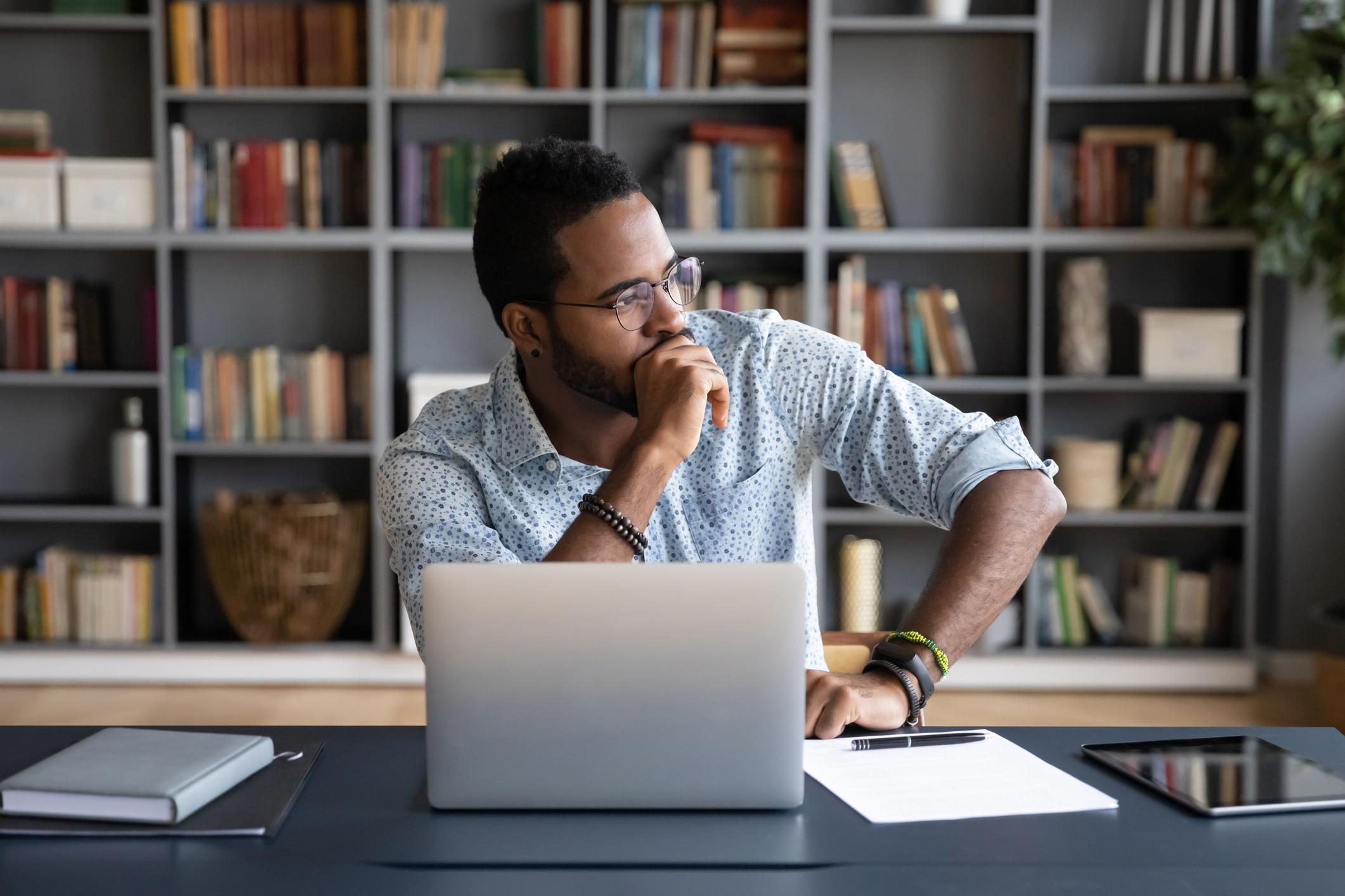 Person sitting at a desk with a laptop, looking thoughtfully out the window in a home office, reflecting on work or personal concerns.