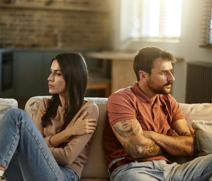 A couple sits apart on a sofa with their arms crossed and faces turned away from each other, suggesting tension or disagreement in their relationship.