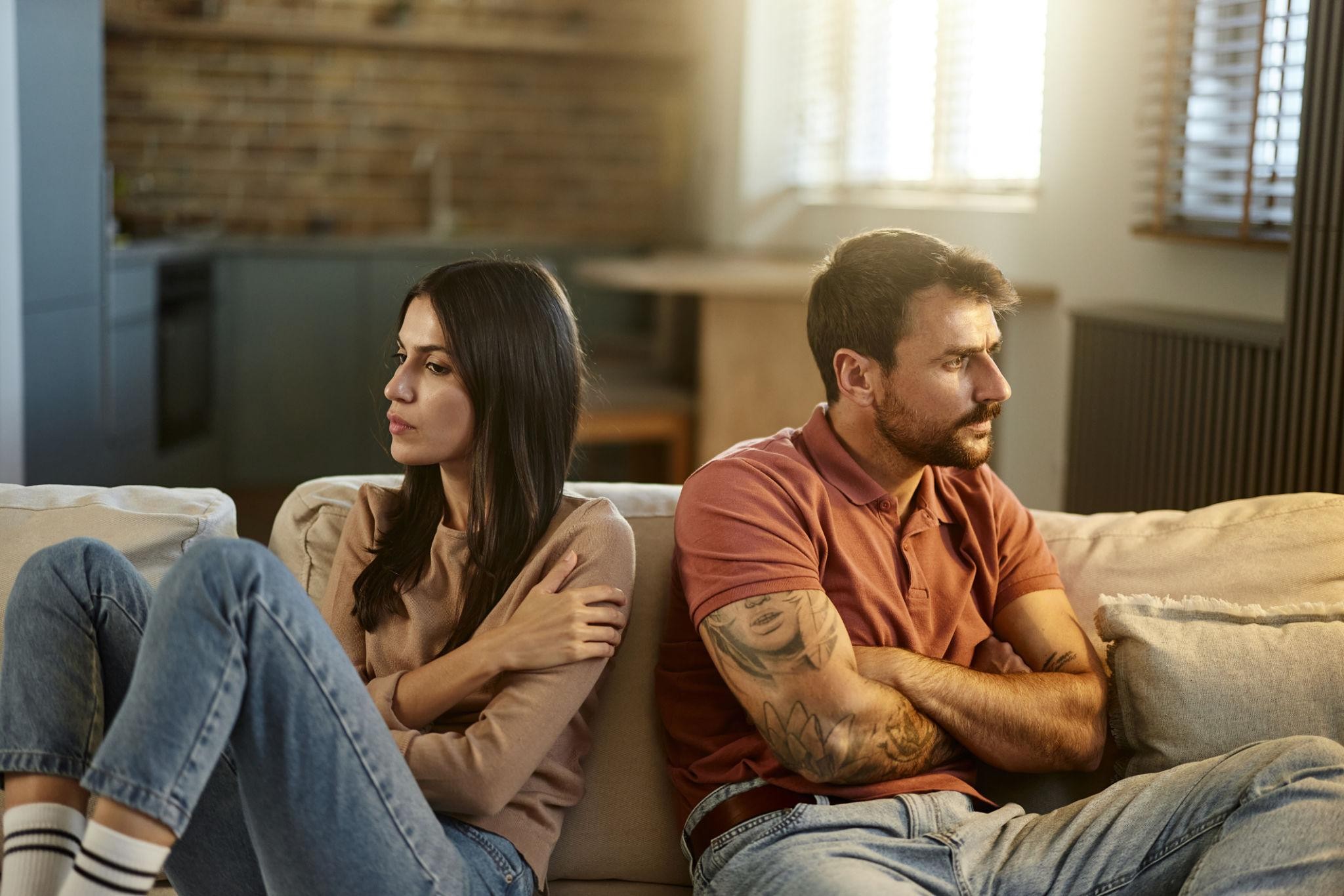 A couple sits apart on a sofa with their arms crossed and faces turned away from each other, suggesting tension or disagreement in their relationship.