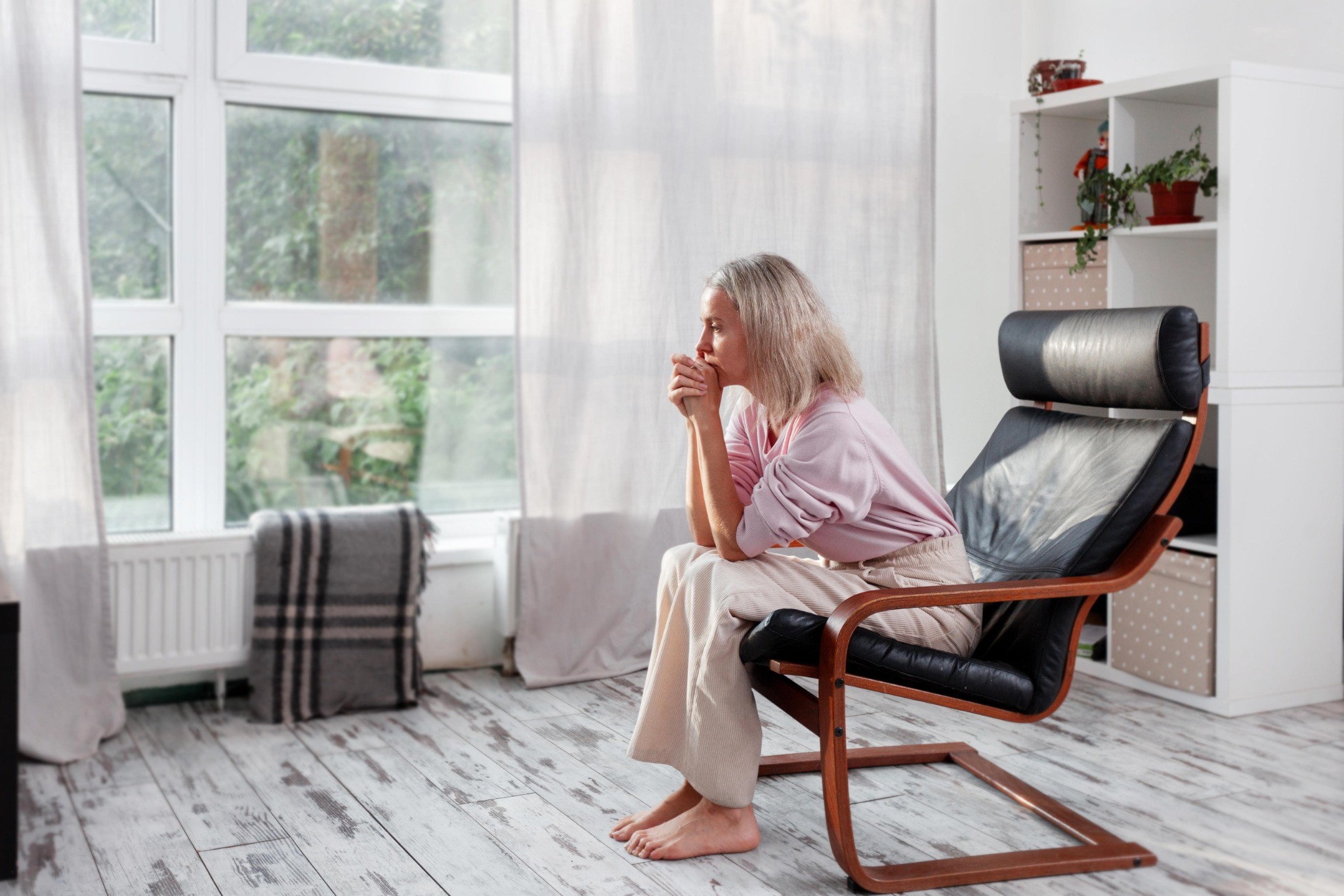An older woman sits alone in a chair by a large window, resting her hands near her face and appearing thoughtful or concerned.
